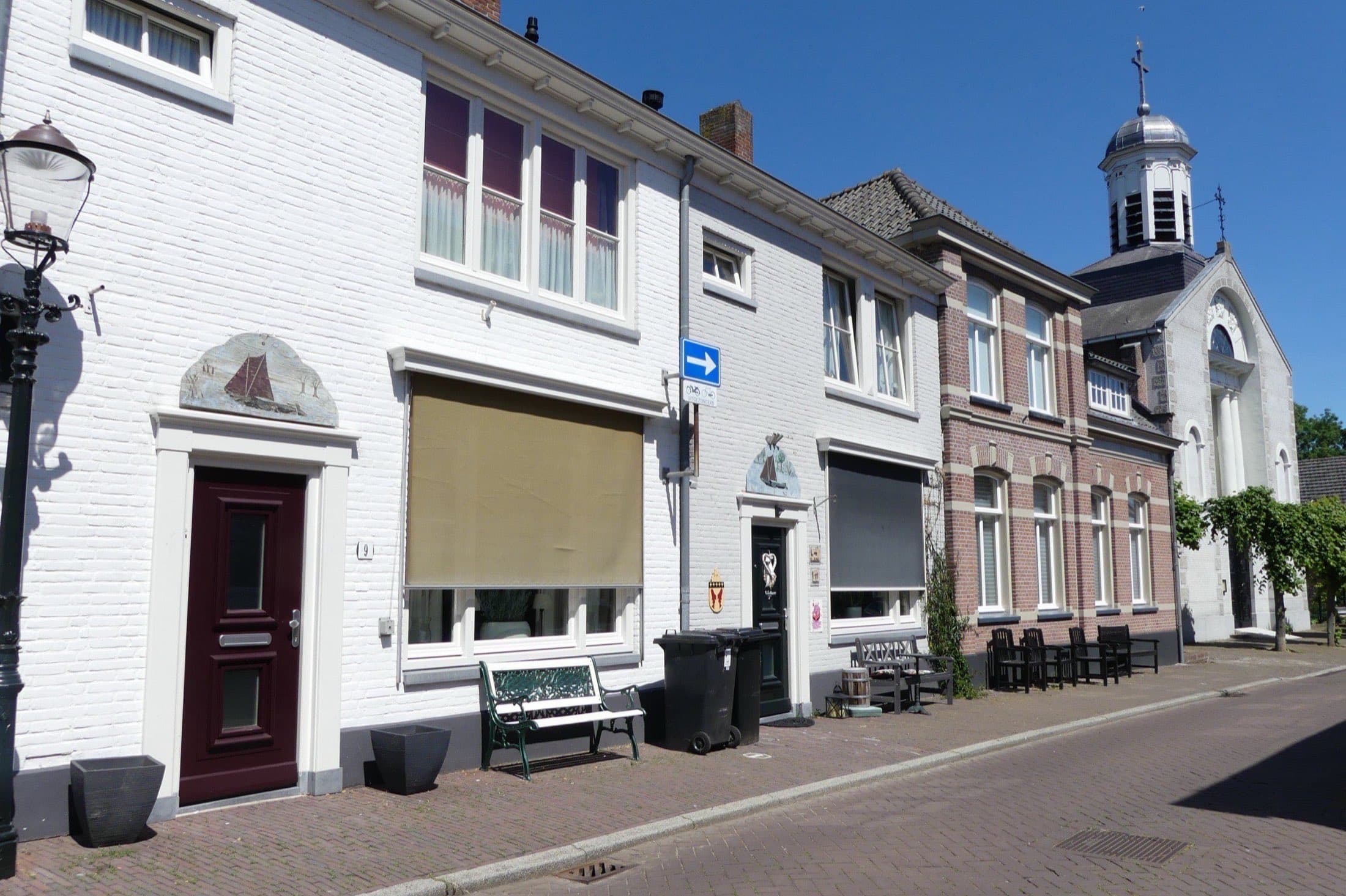 Witgeschilderde huizen aan de Vissersdijk in Woudrichem, gemeente Altena, met op de achtergrond de toren van de Johannes Nepomukkerk onder een blauwe lucht.