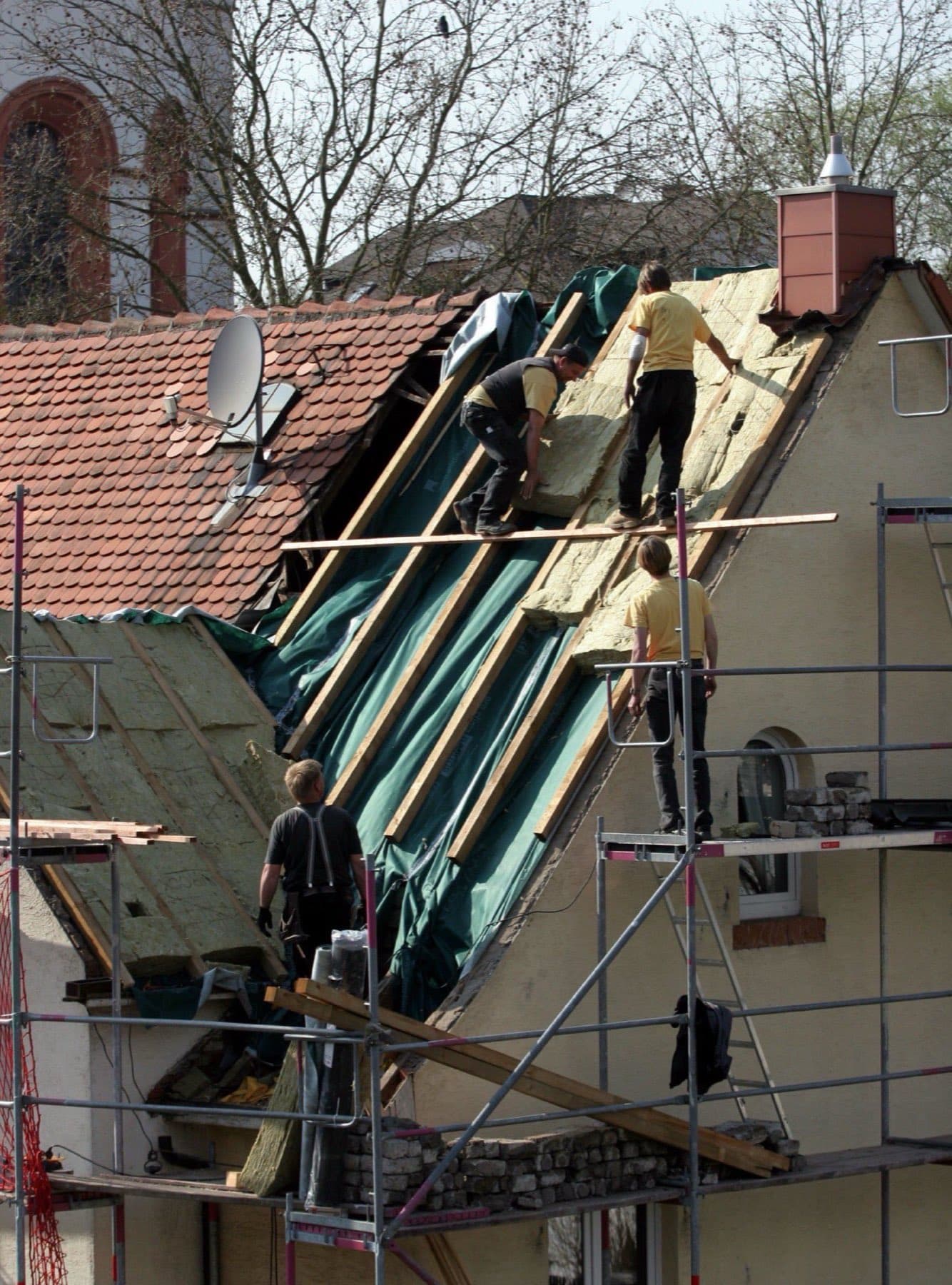 Timmerlieden op een steiger leggen steenwolplaten tussen de dakspanten van een oudere woning. Het dak is aan één kant al open.