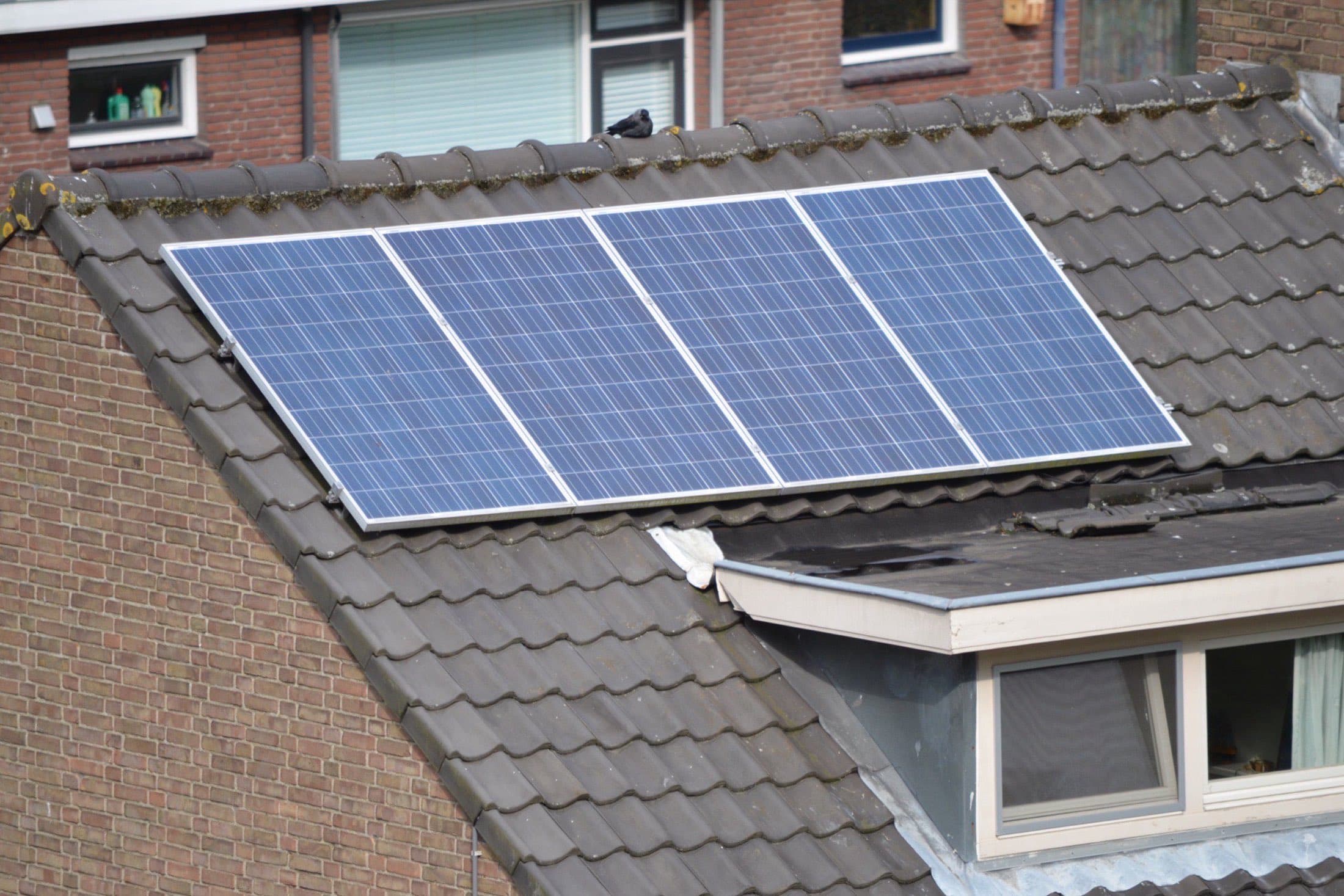 Vier zonnepanelen op een grijs pannendak van een rijtjeshuis, met een dakkapel op de voorgrond.