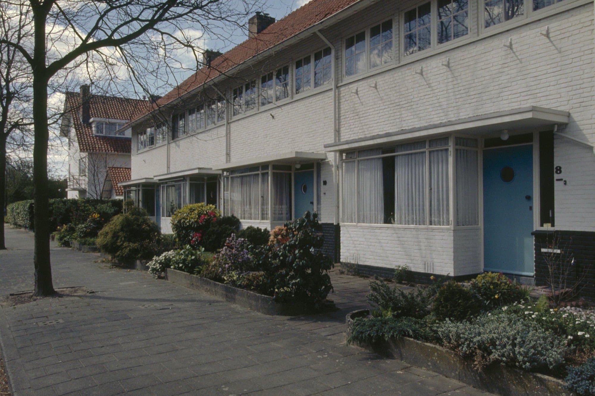 Witte rijtjeshuizen uit de jaren dertig in het Witte Dorp in Eindhoven, met lichtblauwe voordeuren en voortuintjes langs een stoep met bomen.
