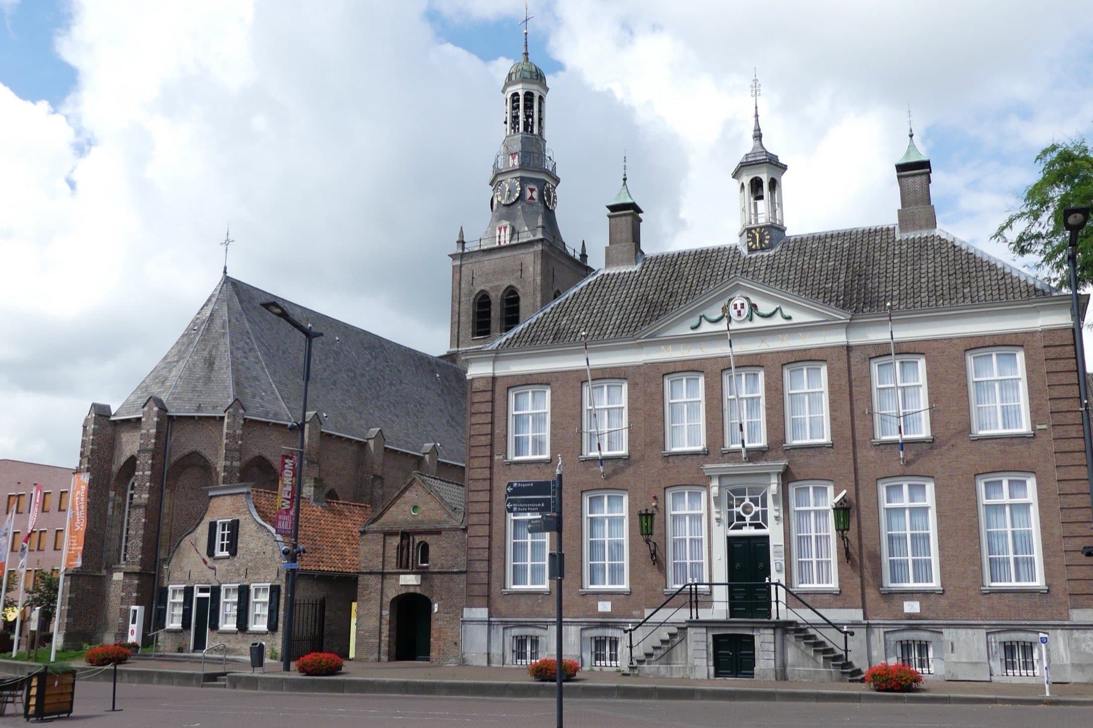 Het oude raadhuis van Etten-Leur met bordestrap en daarnaast de Catharinakerk en een klein huisje aan de Markt, onder een blauwe lucht met wolken.