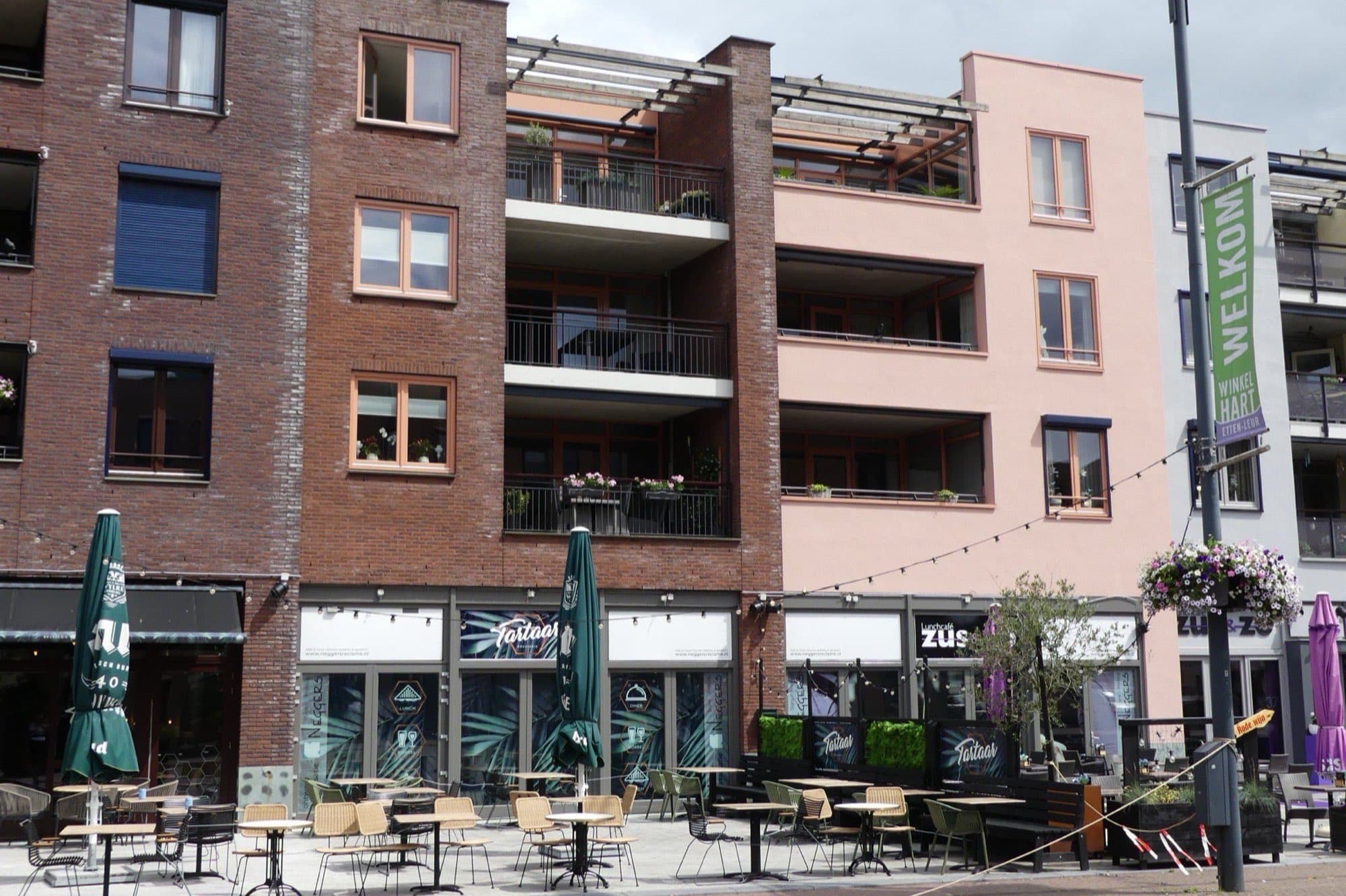 Modern appartementengebouw van donkere baksteen met balkons en een zalmroze zijgevel aan het Raadhuisplein in Etten-Leur, met winkels en een terras op de begane grond.