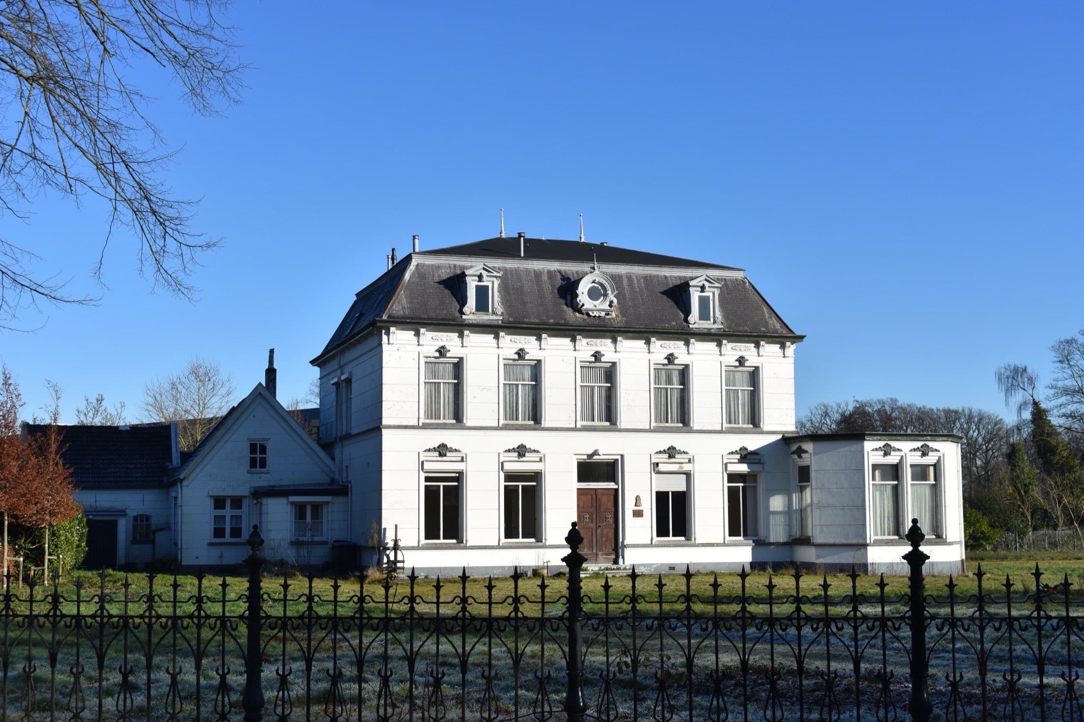 Witgepleisterde villa met een grijs schilddak, dakkapellen en een smeedijzeren hek aan de Postelstraat in Moergestel, gemeente Oisterwijk, onder een blauwe lucht.