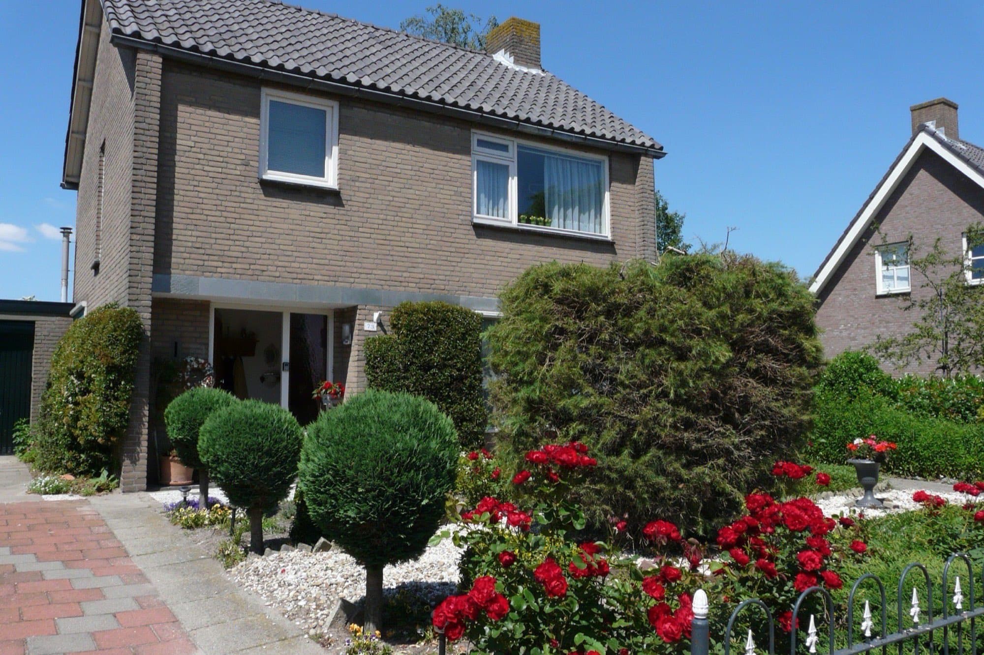 Vrijstaand bakstenen woonhuis met grijs pannendak en een voortuin vol rode rozen en buxusbollen aan de Provincialeweg in Oosteind, gemeente Oosterhout.