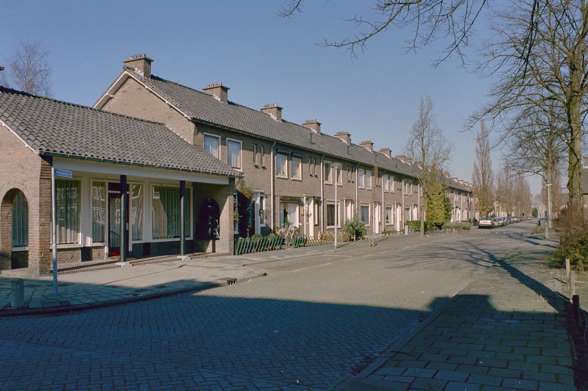 Rij bakstenen rijtjeshuizen met pannendaken en schoorstenen aan de Marconistraat in de Roosendaalse wijk Kalsdonk, met kale bomen op een zonnige dag.
