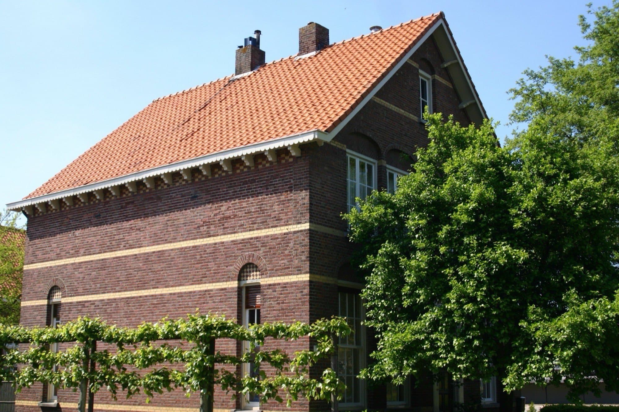 Bakstenen woonhuis met sierbanden, een rood pannendak en een leilinde ervoor aan de Schoolstraat in Wouw, gemeente Roosendaal, onder een blauwe lucht.