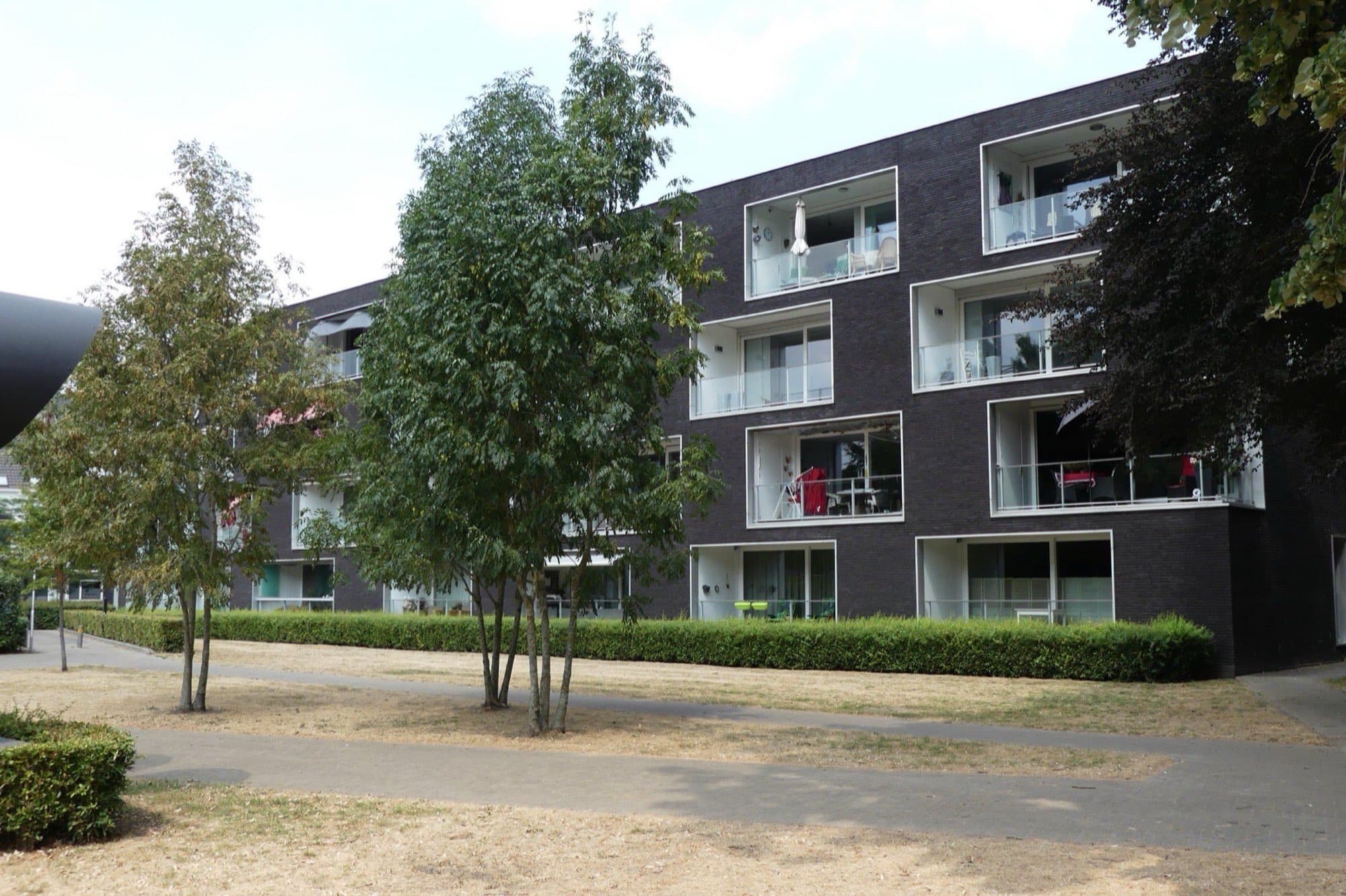 Donker bakstenen appartementengebouw met balkons tussen jonge bomen bij het Harmoniepark in Tilburg.