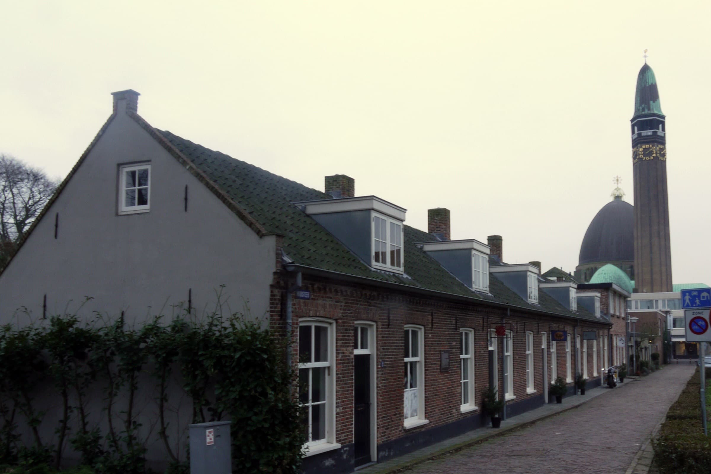 Rij lage witte en bakstenen huisjes met pannendaken aan de Hooisteeg in Waalwijk, met op de achtergrond de toren van de Sint-Jan.